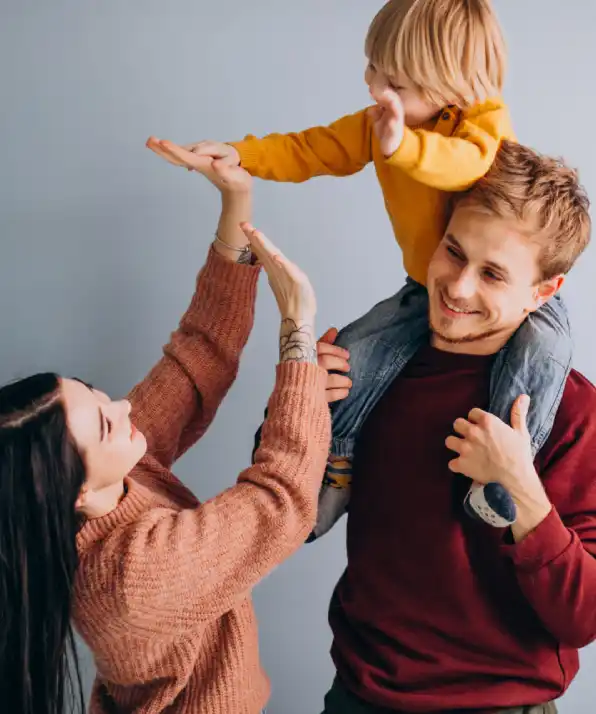 A young child in a yellow shirt is playfully lifted by a person sitting on another's shoulders, against a soft gray background A young child in a yellow shirt is playfully lifted by a person sitting on another's shoulders, against a soft gray background